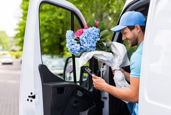A smiling flower courier gets out of a van holding a bouquet of blue and pink flowers. 
