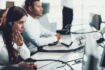 beautiful woman sitting at call center wearing headset talking to client with male colleague in background