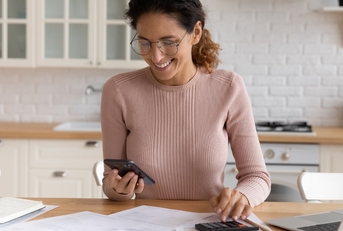 happy woman sorting out budget in kitchen while holding smartphone