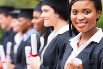 african american female student wearing graduation gown and hat with other students in background at graduation ceremony in college