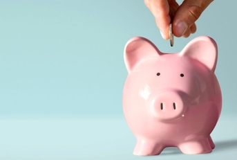 A hand puts a coin into a pink ceramic piggy bank.
