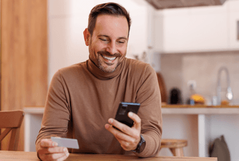 A man in a brown turtleneck smiles while using his phone and holding a credit card in his other hand.