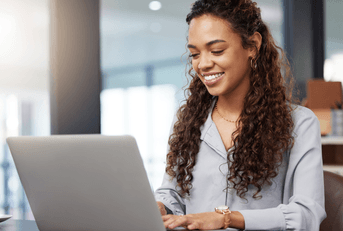 A woman smiles while typing on her laptop in an office setting.