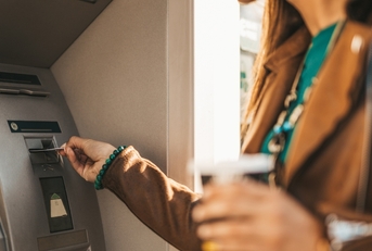 Woman wearing leather jacket and holding a drink using her bank card in an ATM