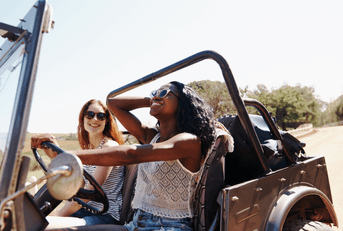 Two women in a convertible SUV drive down a road on a bright, sunny day.