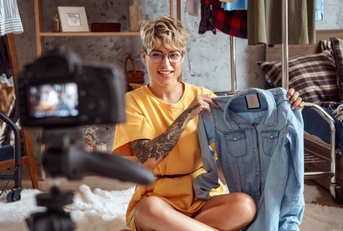 Fashion Blogger. Young woman influencer in glasses sitting on carpet at stylish apartment recording vlog on camera 