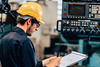 Man wearing yellow protection hat in a factory, taking notes 
