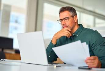 Man in his 40's looking at paperwork and computer thinking