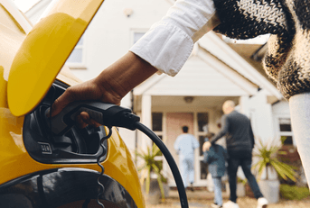 A woman charges a yellow electric vehicle while her family walks into their home in the background.