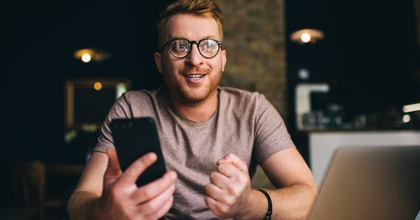 A man wearing glasses sits in front of a laptop and holding a smartphone. 