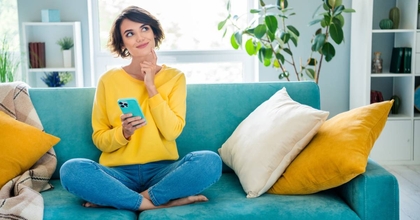 A girl is thinking while sitting on the couch in the living room and holding her phone.