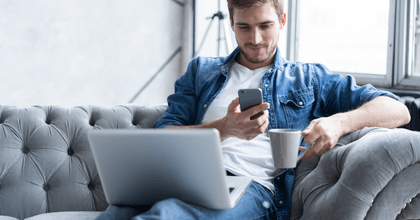 A man sits on the couch and uses his phone and laptop.