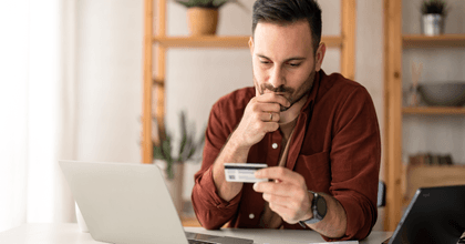 A man appears worried as he holds his chin and looks down at his credit card while sitting in his home office.