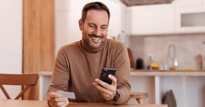 A man in a brown turtleneck smiles while using his phone and holding a credit card in his other hand.