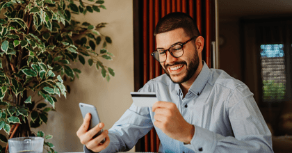 A young man wearing glasses smiles while holding a credit card and looking at his phone.