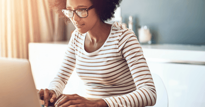 A woman with glasses uses a laptop while sunlight pours through the window.