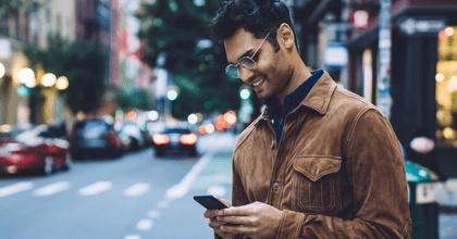 A man wearing glasses stands on the sidewalk in a busy city street while smiling and staring down at his phone.