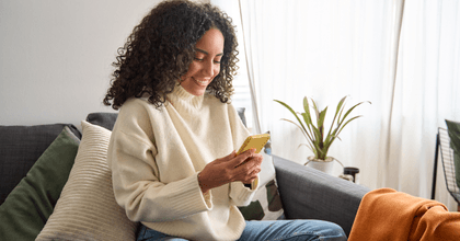 A woman in a creme colored sweater uses her phone while sitting on a gray couch.