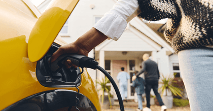 A woman charges a yellow electric vehicle while her family walks into their home in the background.