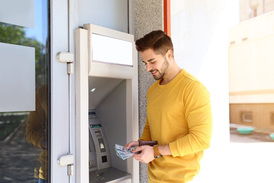 Young man with money near cash machine outdoors