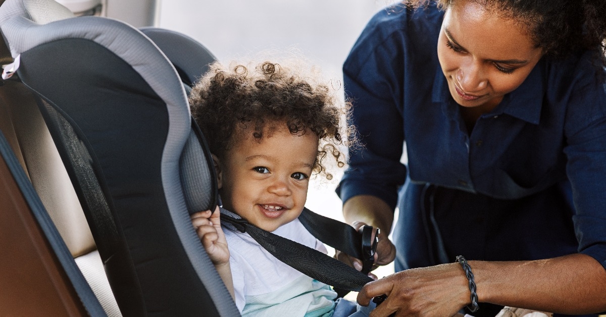 happy little boy looking at camera while his mother buckling him in a car seat