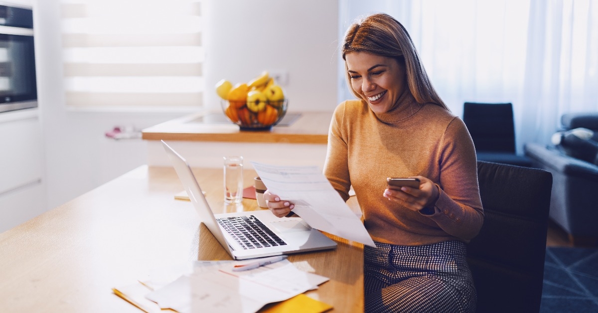  smiling blonde woman in sweater holding bills in one hand and in other smart phone