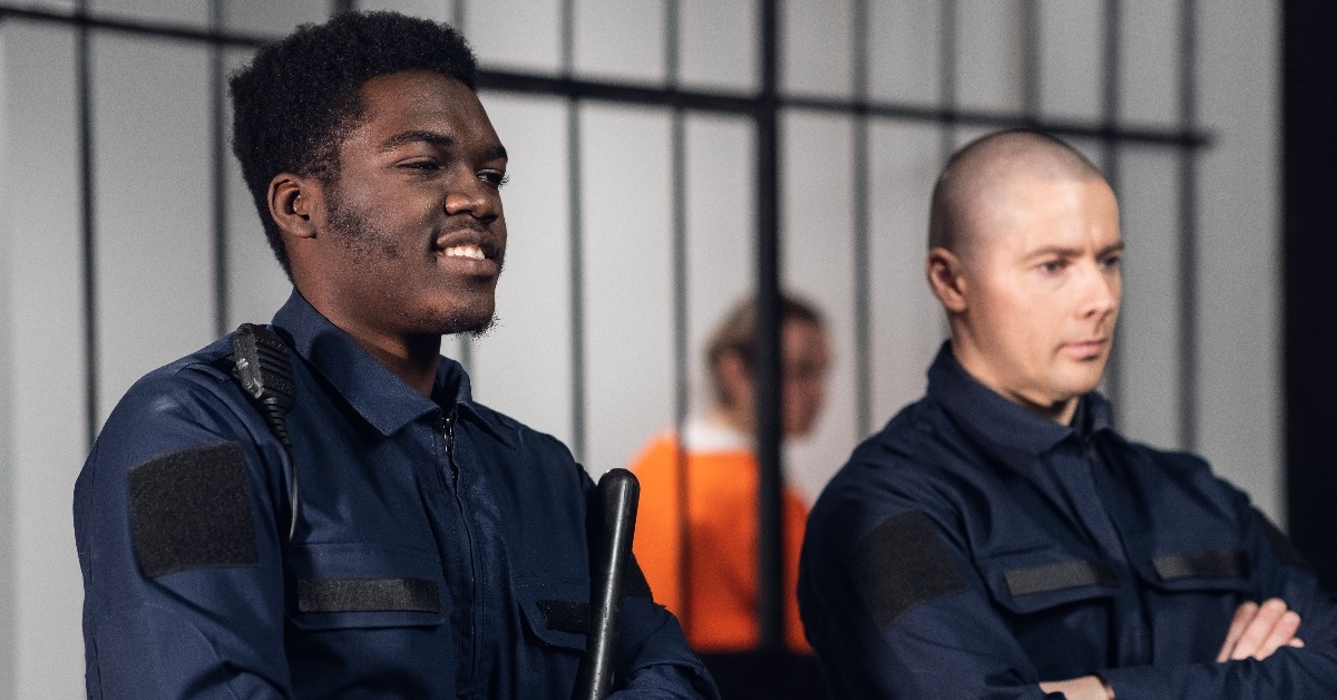 prison guards stand with batons in the background of cells with prisoner