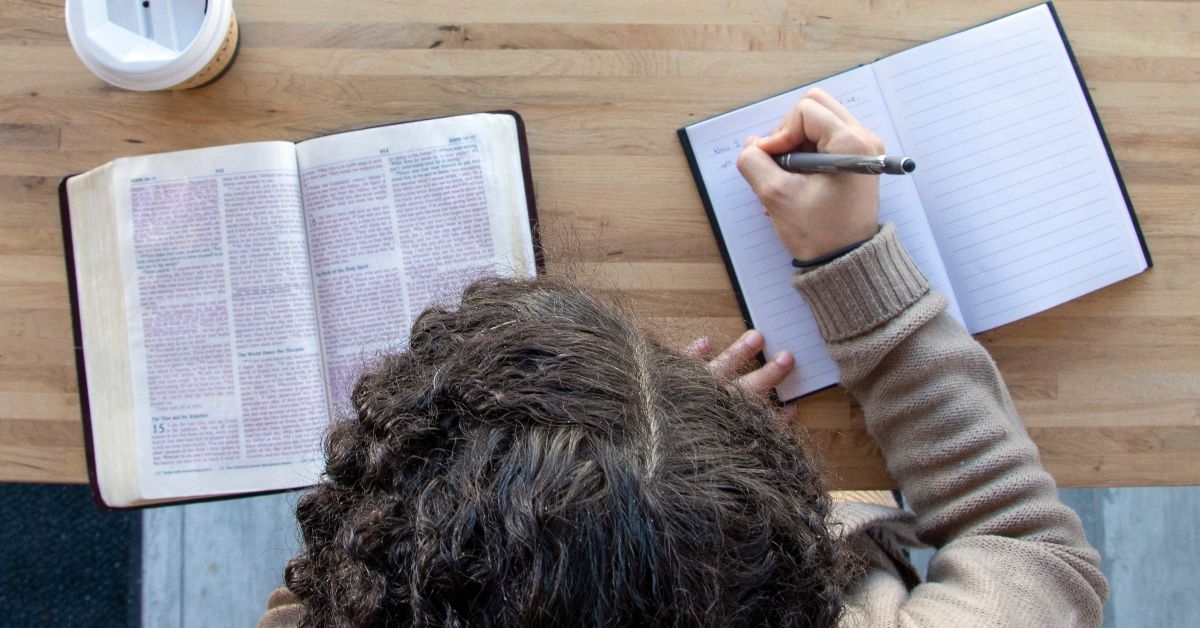 Person writing on notebook while open bible on the side