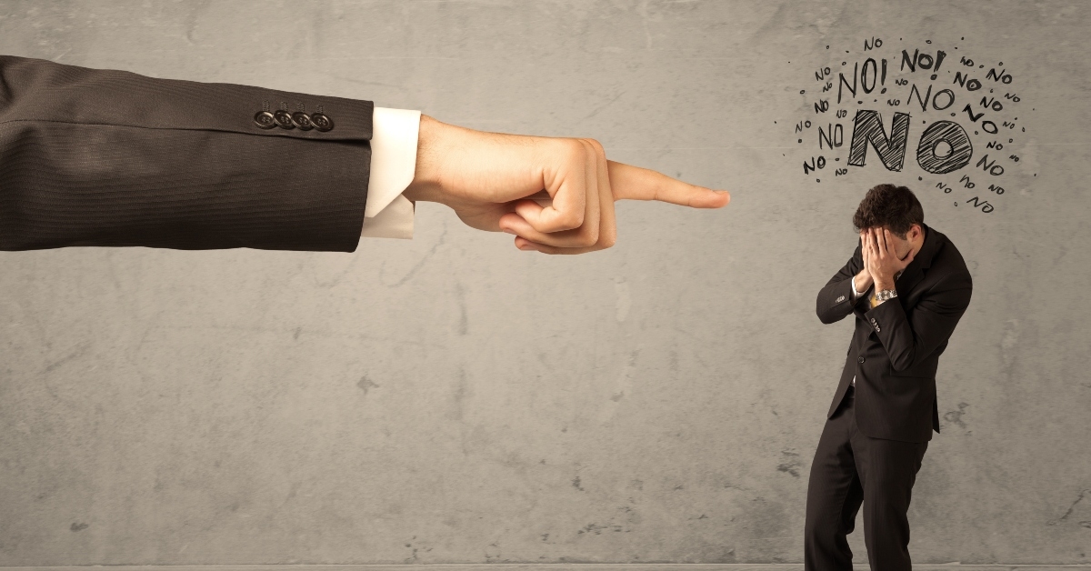 Big hand pointing finger at little man in black suit with hands on his face and many 