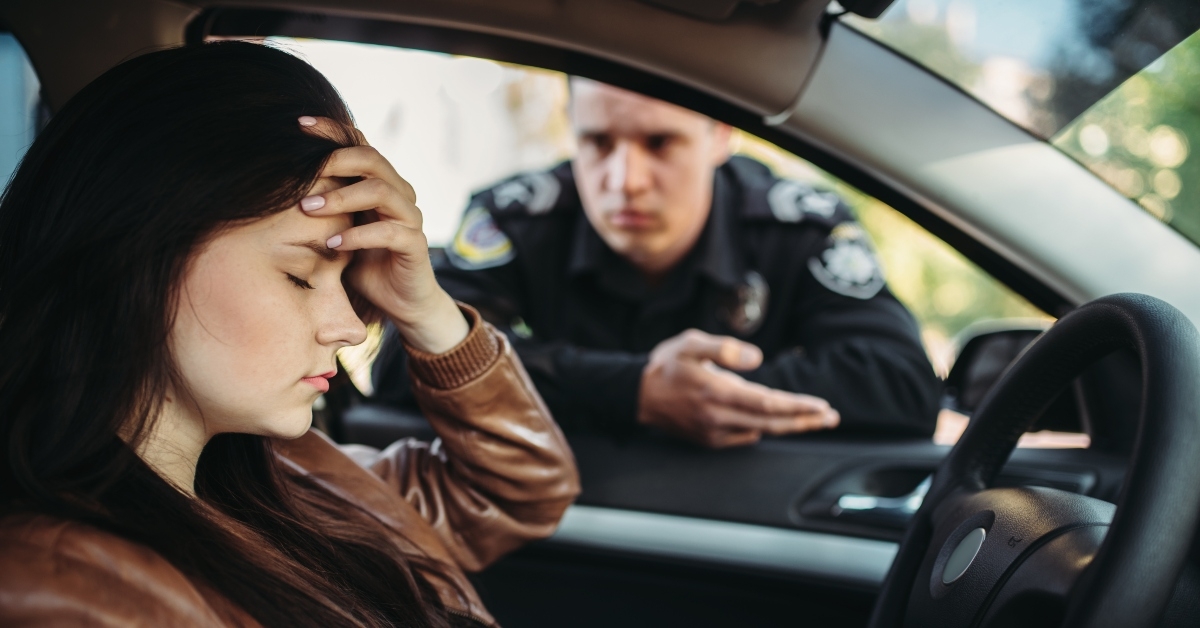 Car parked with young girl driver seat with eyes closed and hand on forehead while police office talks over the window