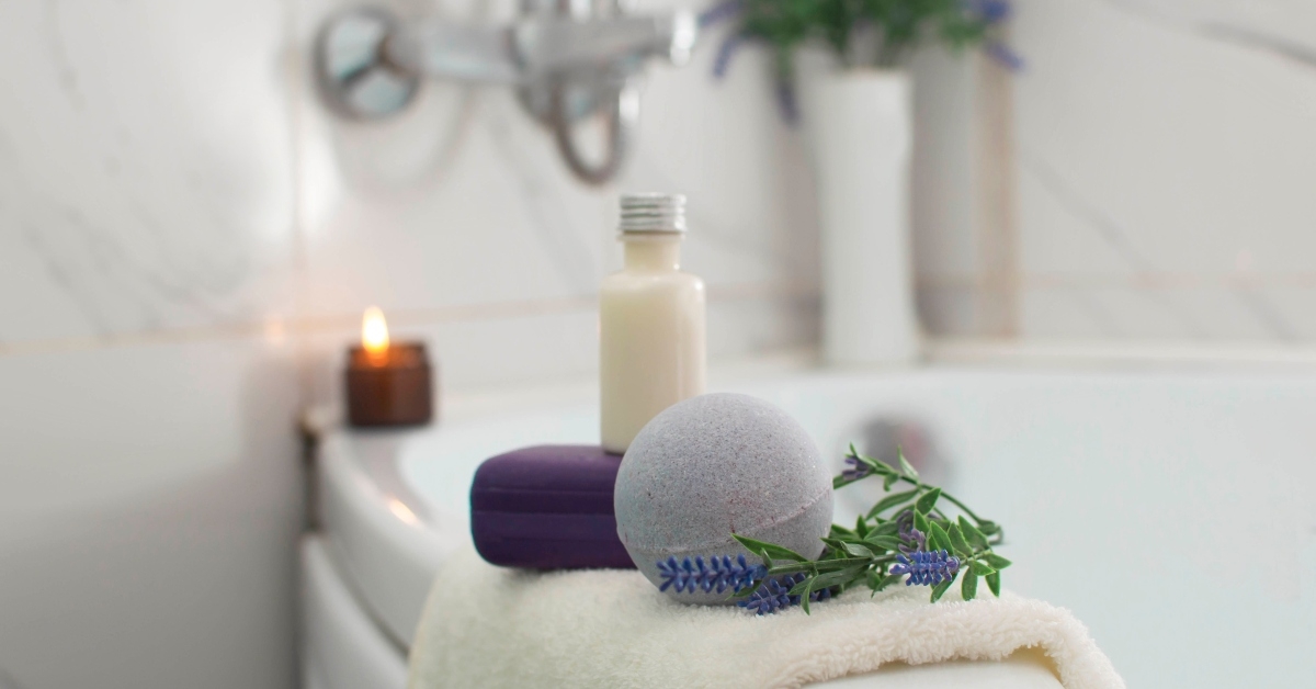 White bathroom and bathtub with candle lit a towel over bathtub with bath bomb, lavender soap and lotion