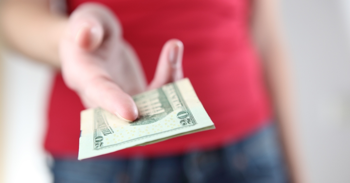 Person in red shirt and jeans blurred in the back with hand reaching in front handing over a twenty dollar bill