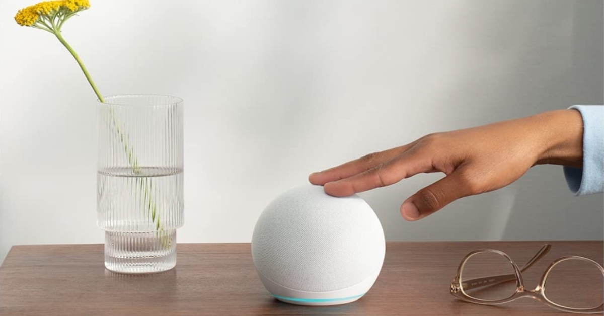 Amazon Echo round white color tech smart home device on wood table with hand touching it next to eyeglass and a glass with water and a yellow flower