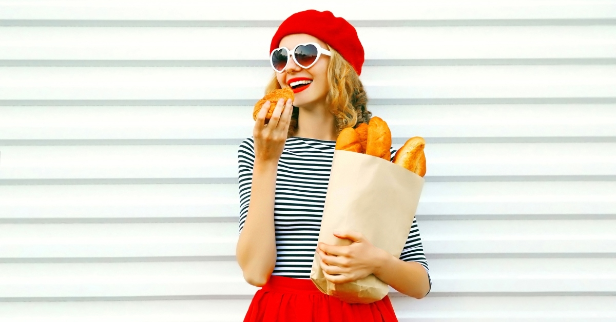 Blonde girl dressed in french style close, red hat, striped shirt, sunglasses, is eating croissant and carrying a bag with baguettes