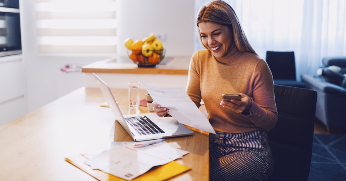 woman sitting on working desk smiling at paper in hand while using smartphone