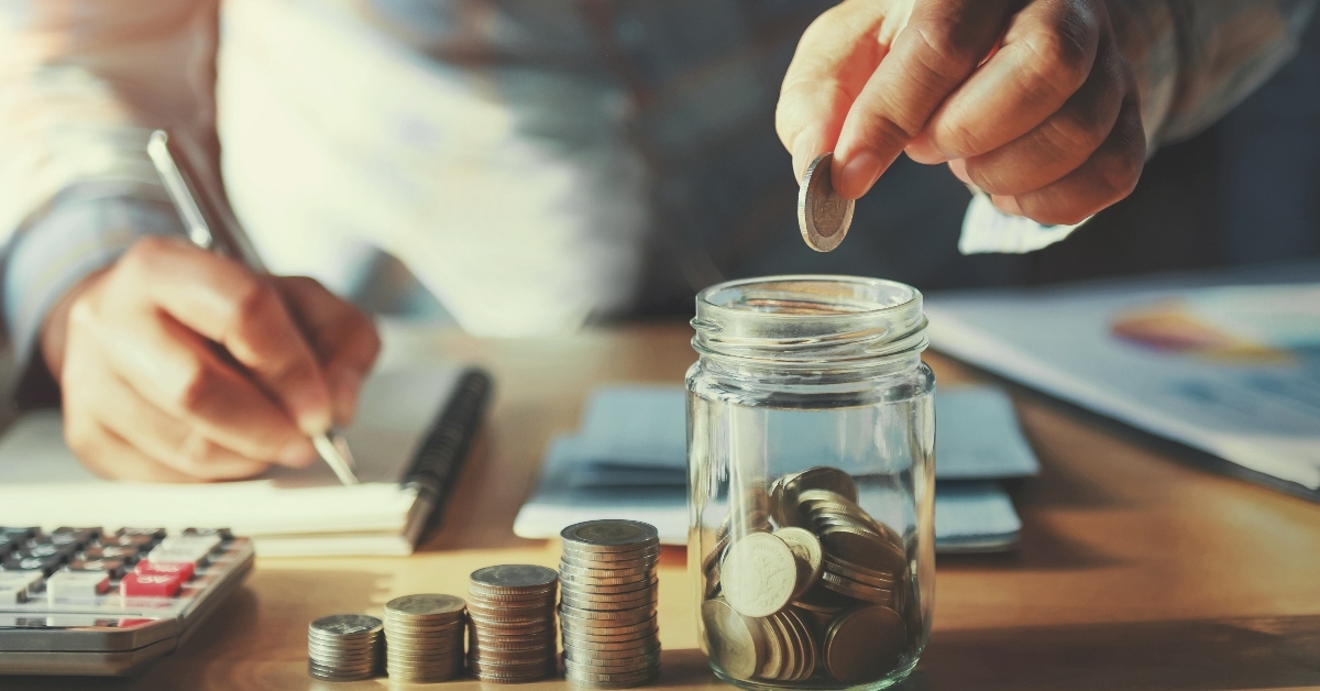 man putting pennies in jar while writing on notebook in background with calculator and penny stack on table