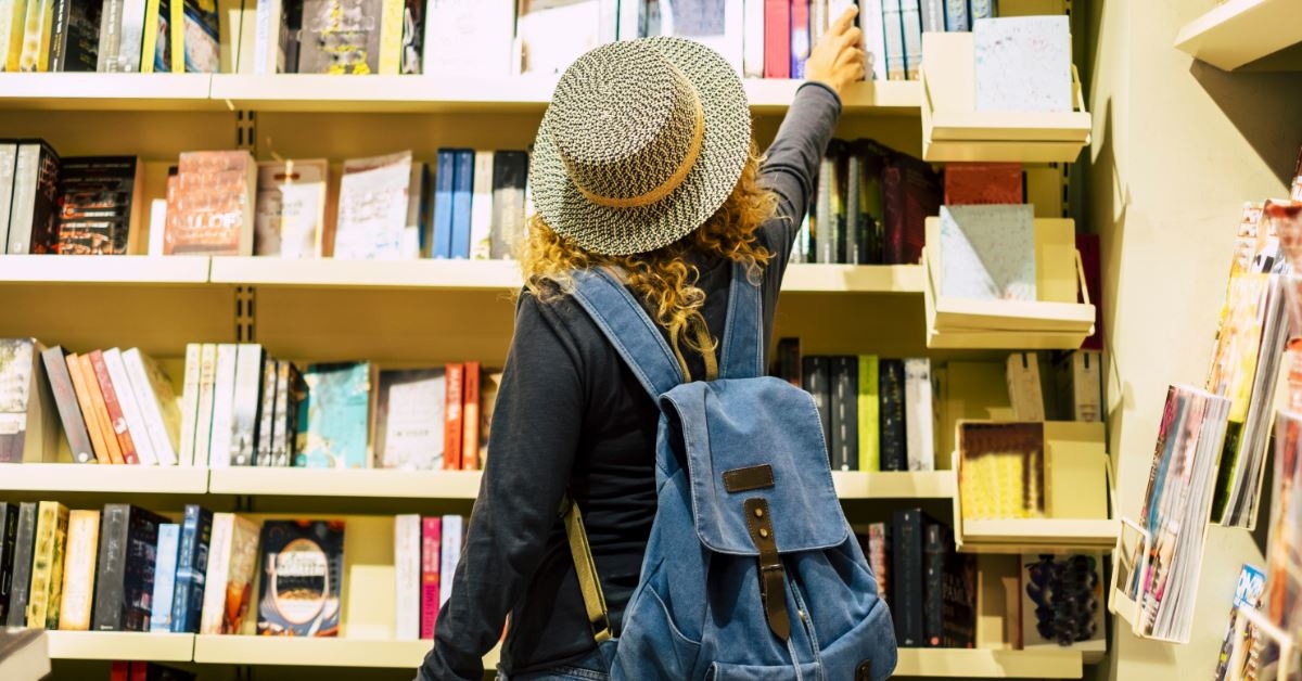young woman with a backpack and hat in library picking out books from shelf