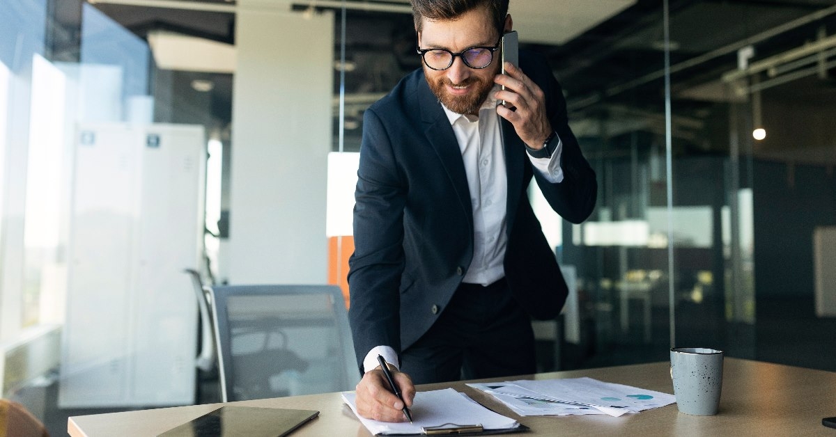 businessman in suit at office talking on cellphone while writing on paper at table
