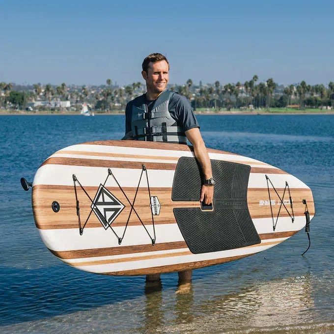 Man holding large white and brown paddle board smiling in front of a blue lake or ocean