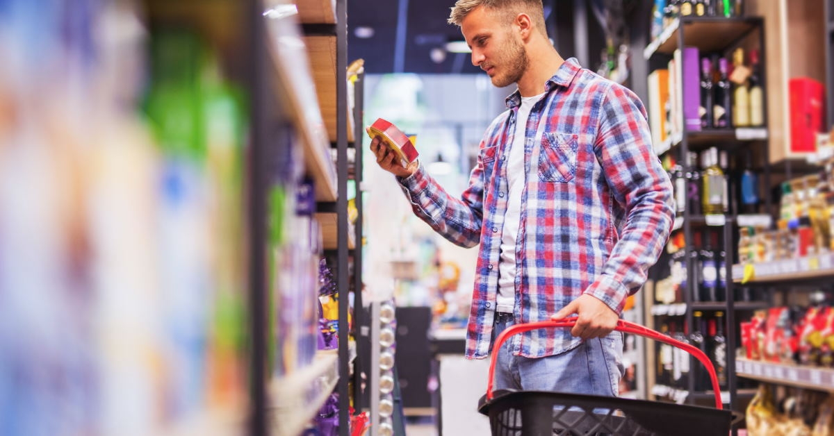 man standing in supermarket buying chocolates from shelf with shopping basket in hand
