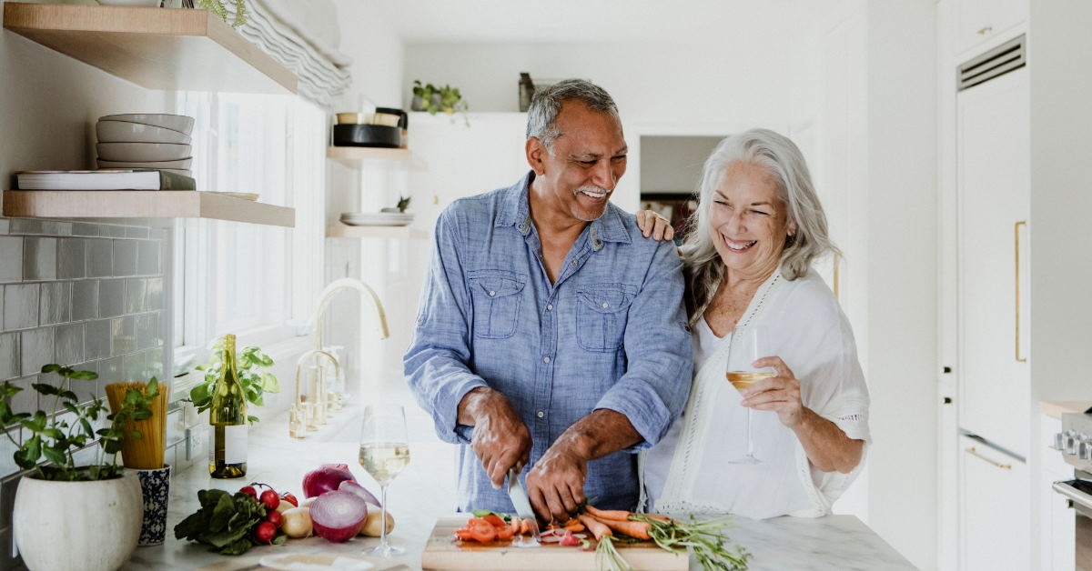 senior couple standing in kitchen at home cutting vegetable holding glass of champagne