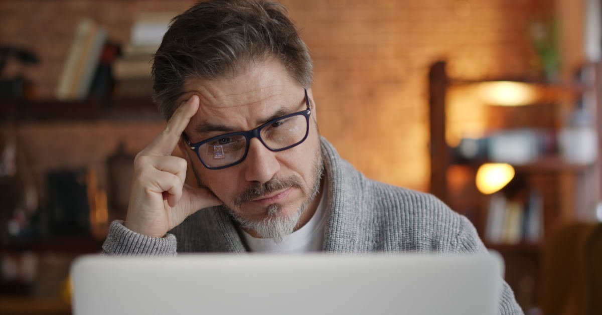 senior man sitting at table using laptop holding head in stress