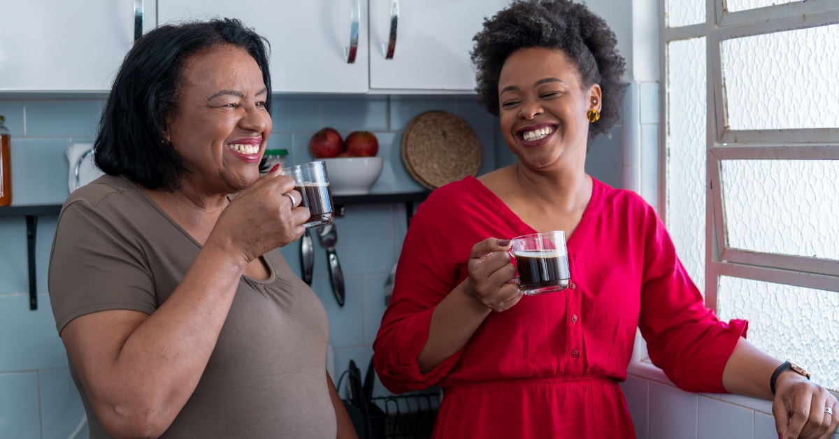 African American senior lady with young lady standing at window holding cup of black coffee at home