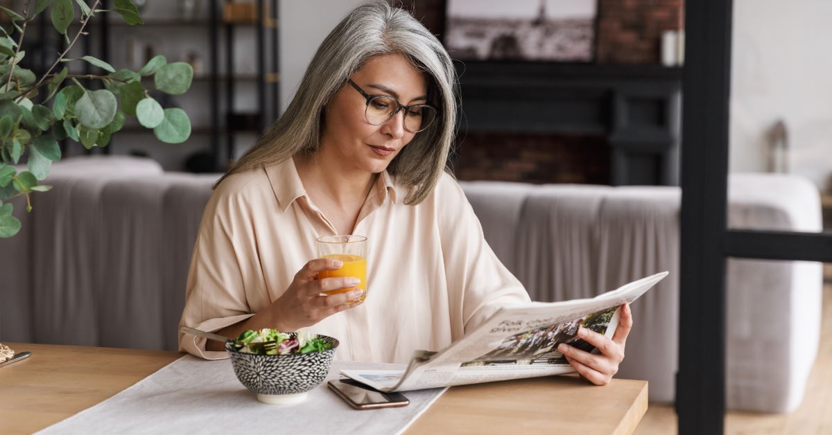 senior woman holding orange juice glass while reading newspaper on table at home