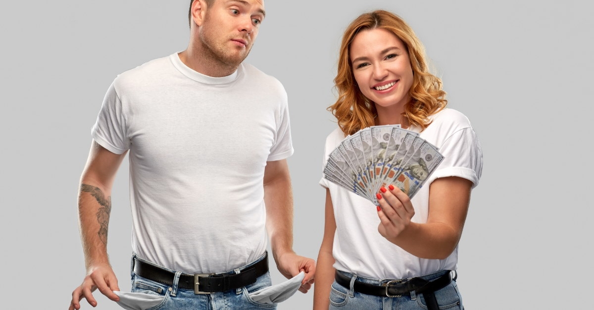 happy woman standing with money in hand with man showing empty pockets on grey background