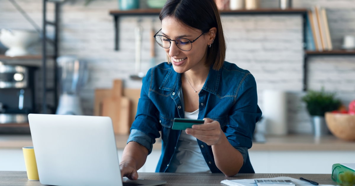 woman sitting at table using laptop while holding card to make online payments
