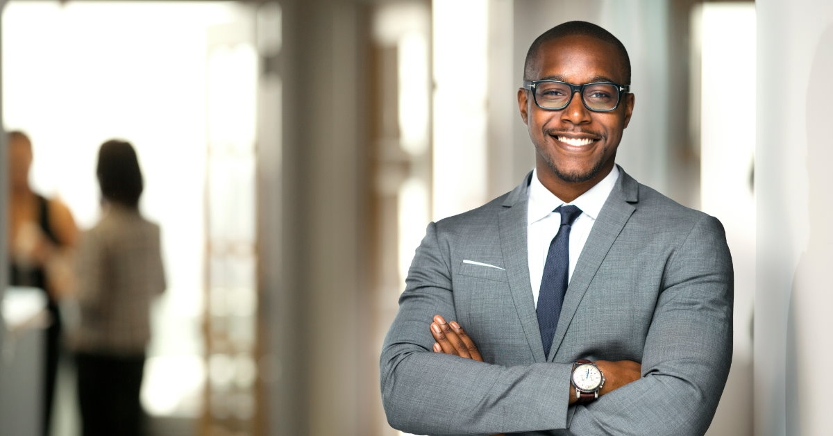 happy african american man in suit standing in office corridor with colleagues in background