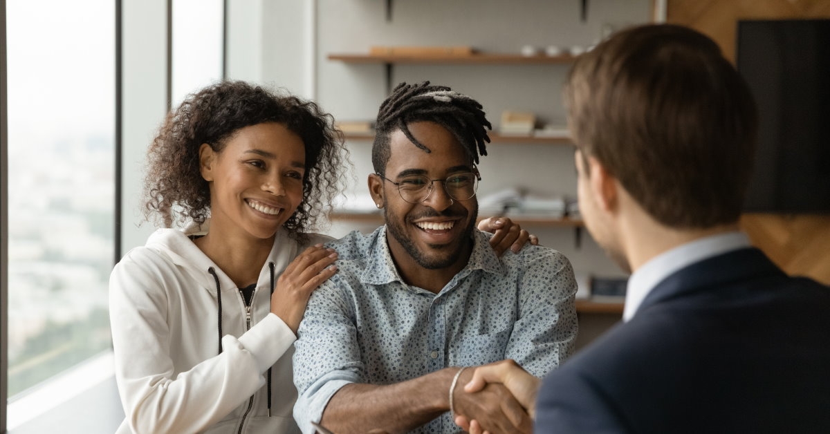 happy couple sitting across table shaking hand with male real estate agent after making deal