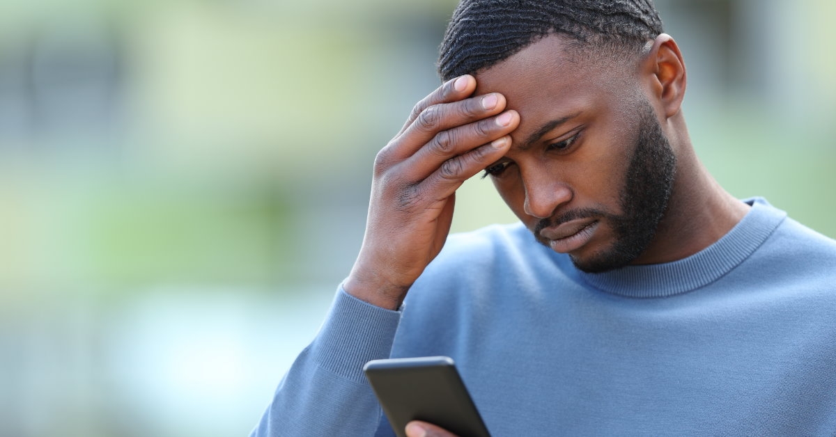 african american man holding his head in shock while using smartphone outdoor