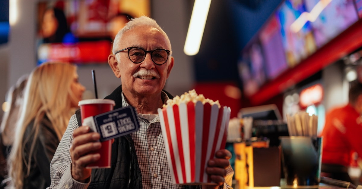 senior man standing in movie theater lounge holding popcorn and drink 